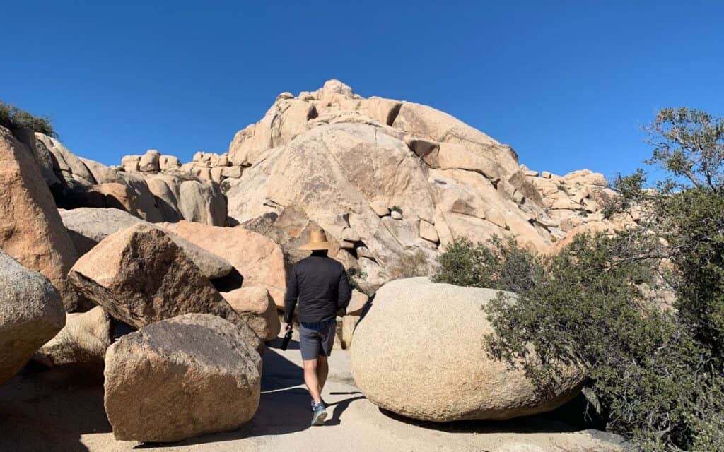 A man walking through two large boulders among large rocks and mountains.