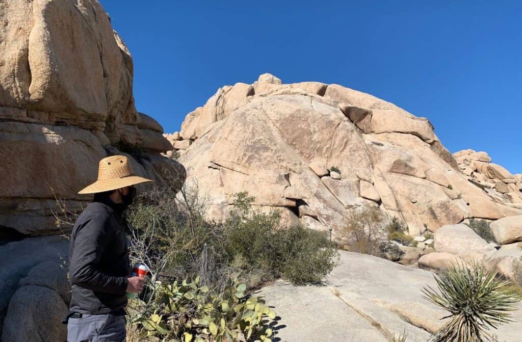 A man walking between boulders with a hat on and holding a cup of coffee.