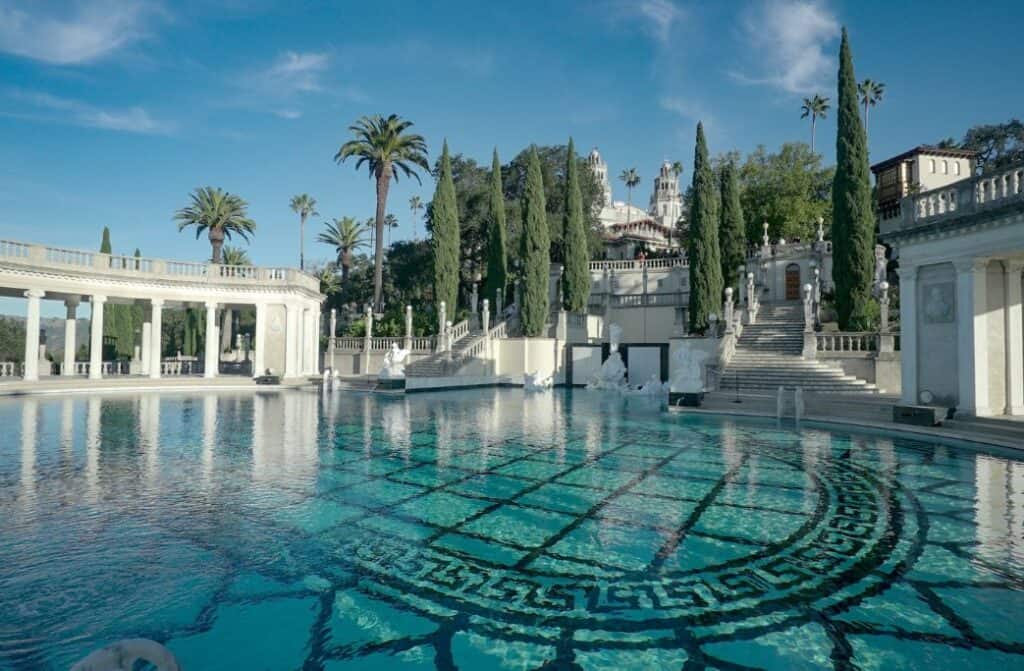 A pool surrounded by greek architecture and tll green trees.