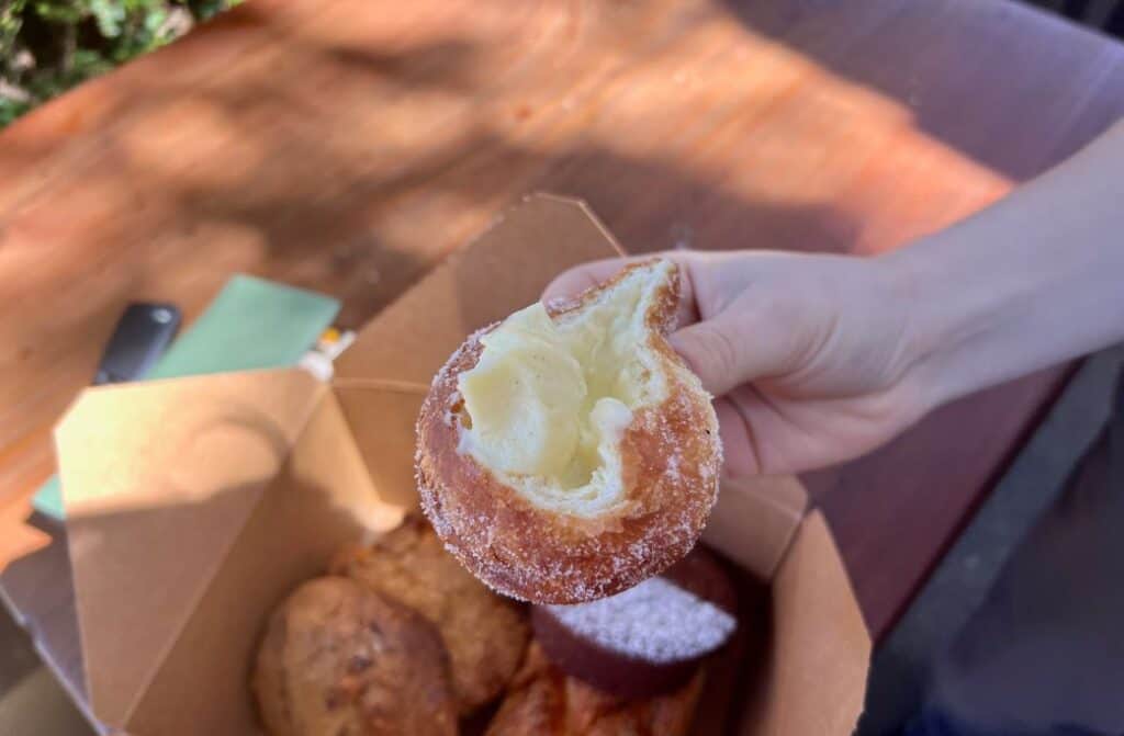 A hand holding a cream-filled donut over a box of pastries.