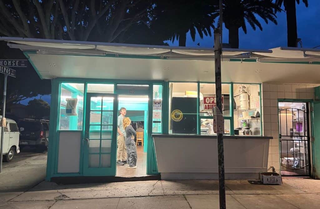 A man and a girl stand in a taco stand at night with palm trees overhead.