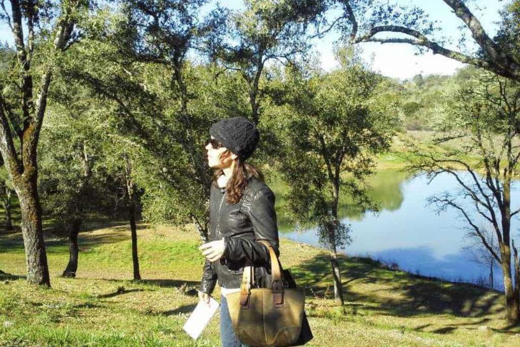 A woman in a black hat and black leather jacket holding a purse in front of a lake and trees in Napa Valley in the fall.