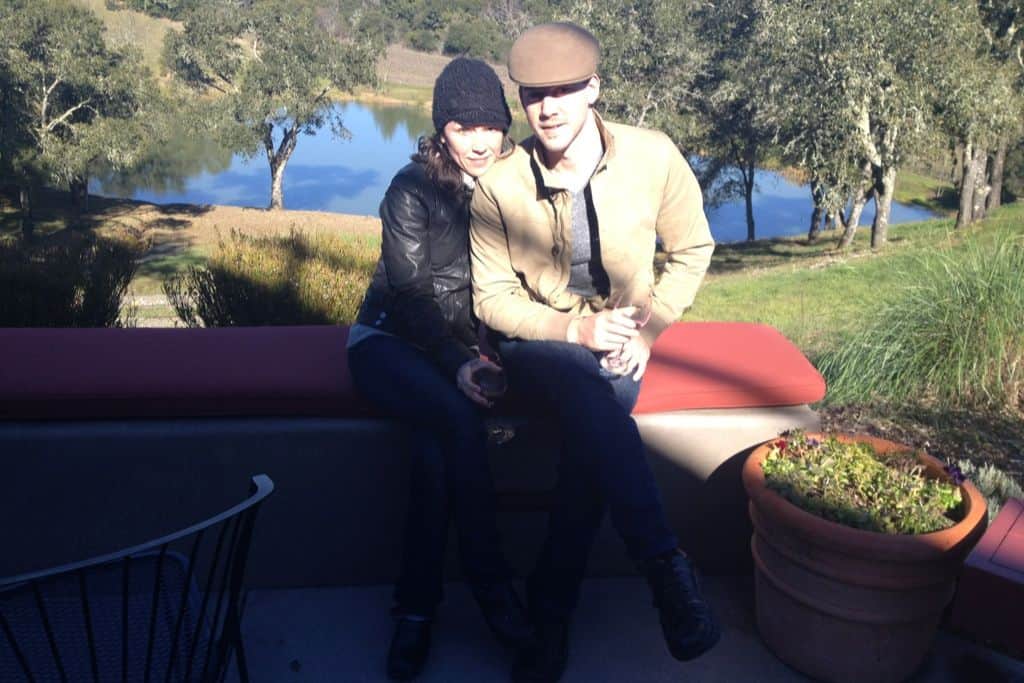 A handsome man and a woman sit on a bench with a lake behind them.