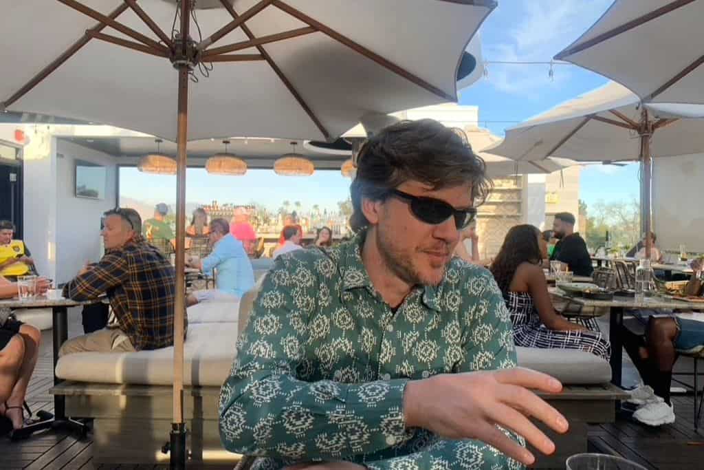 a man in sunglasses reaches for his drink at the table. He is sitting in an outdoor restaurant with people behind him having a good time.