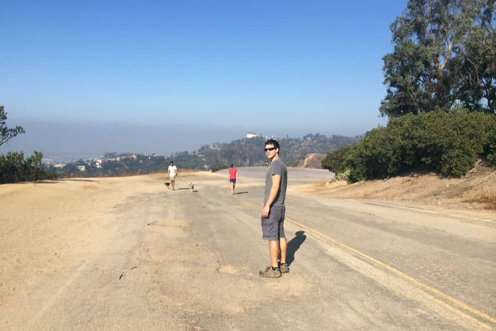 a man looking at the camera on a hike on a gravel path with hills and the Griffith Observatory in the background.
