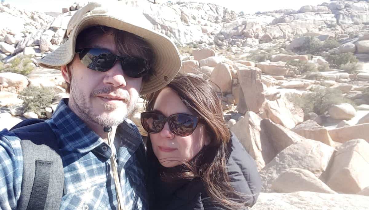 A hot man and woman pose in front of large boulders on the Hidden Valley hike Joshua Tree National Park.
